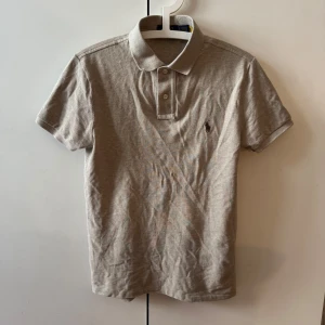 Beige pikétröja Polo Ralph Lauren - Klassisk beige pikétröja från Polo Ralph Lauren i slim fit-modell. Tröjan har korta ärmar, krage och knappar framtill samt den ikoniska broderade loggan på bröstet. Tillverkad i mjuk bomull som känns skön mot huden.
