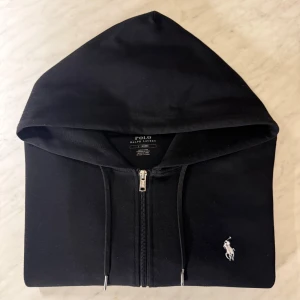 Ralph Lauren zip hoodie - En svart Ralph Lauren tröja med en vit logga ✔️ med en dragkedja tröjan är i storlek L.                            Mått 📐 (midja 91-94 cm) (höft 103-106 cm) Ovan byst 104-107 cm. För fler bilder eller frågor om storlek kontakta mig så får ni svar 💛