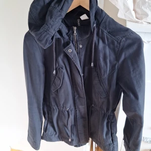 Svart parkajacka från Divided, strl 38 - Svart parkajacka från Divided med huva och dragsko i midjan. Jackan har dragkedja och knappar framtill, samt praktiska fickor med lock. Perfekt för dig som gillar en enkel och stilren look. Långärmad och i mjukt bomullsmaterial.