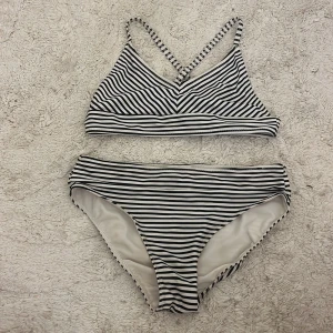 Randig bikini från H&M, svartvit - Snygg bikini från H&M i svartvitt randigt mönster. Överdelen har smala axelband som korsas i ryggen och byxorna har klassisk passform. Perfekt för stranden eller poolen, enkel och stilren design som alltid funkar.
