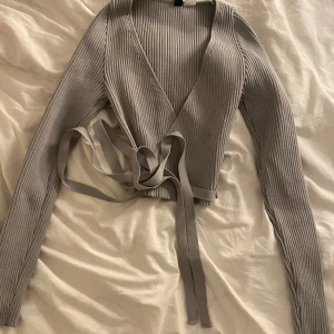 Grå ribbad omlottkofta från H&M Divided - Supersnygg grå ribbad omlottkofta från H&M Divided i storlek XS. Tröjan har långa ärmar, v-ringning och knytband i midjan för en tight och trendig passform. 