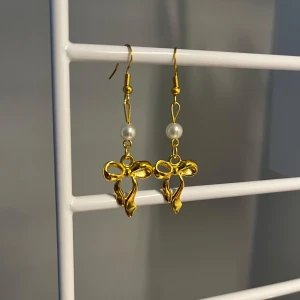 Guldiga örhängen med rosett och pärla - Snygga örhängen i guldton med hängande rosettformad detalj och en liten vit pärla ovanför. Perfekt accessoar för att lyfta din look med en söt och trendig touch. Handgjort 