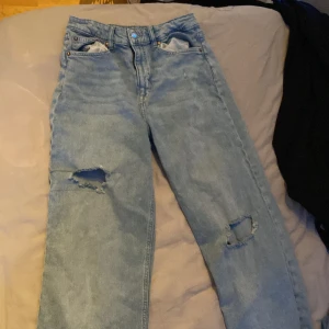 Ljusblå ripped jeans med raka ben - Säljer ett par ljusblå jeans med raka ben och snygga slitningar på båda knäna. Klassisk femficksmodell med hög midja och råa benslut. Perfekta för en avslappnad streetstil och tillverkade i äkta denim.
