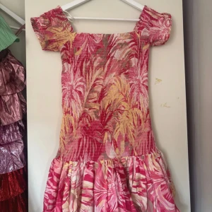 Rosa och gul blommig kortklänning - Superfin kortklänning med tropiskt blommigt mönster i rosa, gult och beige. Klänningen har korta ärmar, smockad överdel och volangig kjol. Perfekt för sommarens alla tillfällen och ger en färgglad vibe.