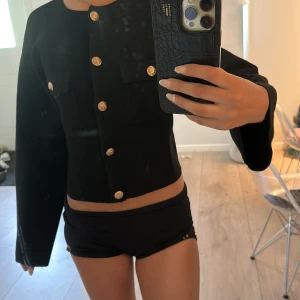 Svart kofta/jacka  med guldknappar - Stilren svart kofta med raka axlar och croppad passform. Jackan har dekorativa guldknappar framtill och på fickorna, samt två bröstfickor. Perfekt för dig som vill ha en klassisk men edgy look.