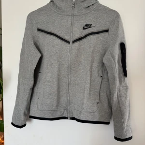 Grå zip-hoodie från Nike - Grå zip-hoodie från Nike med svarta detaljer och logga på bröstet. Jackan har huva, hel dragkedja och en praktisk ficka med dragkedja på ärmen. Tillverkad i mjukt bomullsmaterial, perfekt för chill dagar eller träning.