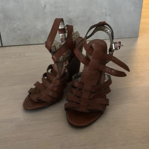 Bruna kilcklacksandaler i skinn  - Bruna sandaler med kilklack och öppen tå. Remmar i mjukt skinn och spänne runt ankeln. Perfekta för dig som vill ha både höjd och komfort. Klackarna har trälook. 