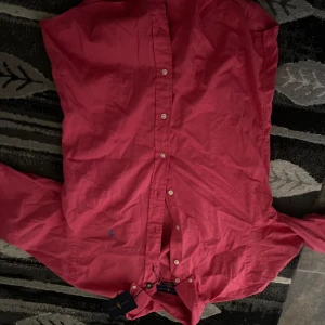 Röd skjorta från Ralph Lauren - Snygg röd skjorta från Ralph Lauren med klassisk krage och knäppning framtill. Skjortan har lång ärm och är tillverkad i mjuk bomull. På bröstet finns det ikoniska broderade Polo-loggan i blått. Perfekt för dig som vill ha en stilren och tidlös look.