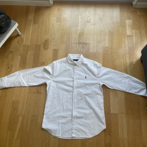 Vit skjorta från Ralph Lauren - Klassisk vit skjorta från Ralph Lauren med blå broderad logga på bröstet. Skjortan har lång ärm, knappar hela vägen och en snygg krage. Tillverkad i bomull för en skön och stilren känsla. Perfekt för dig som gillar enkel och tidlös stil. Köpt för ett tag sen men har inte fått någon användning pga att den är lite för stor för mig. Den är nyskick och det är bara att höra av sig vid frågor