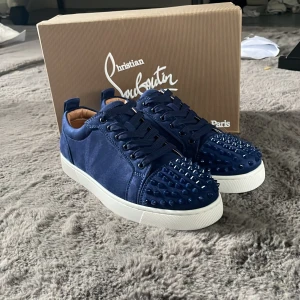 Blå Christian Louboutin sneakers med nitar - Snygga blå sneakers från Christian Louboutin med coola nitar på tån och klassisk röd sula. Skorna har snörning, vit platt sula och är tillverkade i ett lyxigt material som ger en exklusiv känsla. Perfekta för dig som vill sticka ut med din stil.