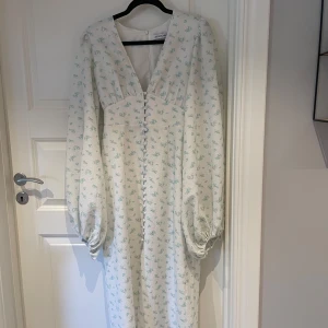 Vit blommig långklänning från Adoore - Superfin långklänning från Adoore i vitt med ljusblått blommönster. Klänningen har v-ringning, långa puffärmar och knappar framtill. Perfekt för sommarens alla tillfällen och ger en romantisk vibe.