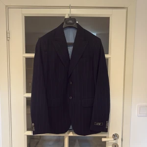 Mörkblå randig kostym från SIR - Snygg mörkblå kostym från SIR of Sweden med tunna ljusa ränder. Kavajen har klassisk passform, breda slag och två knappar framtill. Byxorna är raka med pressveck och uppvikta benslut. Perfekt för dig som vill sticka ut med stilren look. Bara bärd 1 gång 