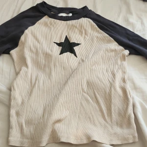 Beige och svart långärmad tröja H&M - Ribbad långärmad tröja från H&M i beige med svarta ärmar och svart stjärna på bröstet. Skön passform och cool kontrast mellan färgerna. Perfekt för dig som gillar en enkel men ändå snygg stil. Den är kort (det är meningen) som en crop top