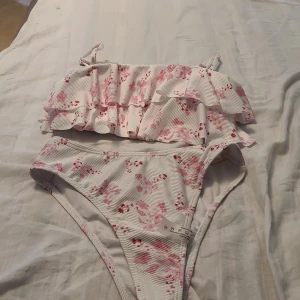 Vit blommig bikini med volang - Supersöt bikini i vitt med rosa och röda blommor. Toppen har volangdetaljer och smala axelband, medan bikinitrosan har hög midja. Materialet är ribbat och stretchigt, perfekt för strandhäng. Säljs som set.