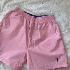 Rosa badshorts från Polo Ralph Lauren - Snygga rosa badshorts från Polo Ralph Lauren med elastisk midja och innerfoder i mesh. Klassisk blå logga broderad framtill och praktisk bakficka med lock. Perfekta för stranden eller poolen i sommar. Storlek M ’en passar även S. Aldrig använda för små.