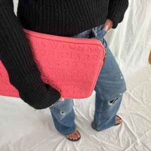 Rosa datorväska från Marc Jacobs - Säljer en snygg rosa datorväska från Marc Jacobs med präglad logotyp över hela väskan. Väskan har en rektangulär form, dragkedja i guld och är tillverkad i ett mjukt syntetmaterial. Perfekt för att bära din laptop med stil.