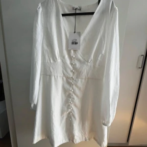 Vit kortklänning med knappar - Superfin vit kortklänning med v-ringning och långa ärmar. Klänningen har dekorativa knappar framtill och lätt puffade axlar. Tillverkad i ett tunt, lätt material som ger en luftig känsla. Perfekt för en fräsch och stilren look. Prislappen är ej kvar 