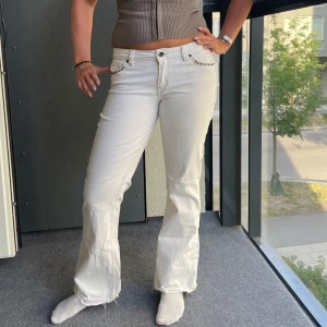 Vita bootcut jeans med detaljer - Säljer ett par vita bootcut jeans med snygga nitar och broderier på fickorna. Jeansen har låg midja och råa kanter nertill för en cool vibe. Perfekta för dig som vill sticka ut lite extra. Materialet är bomull och de har klassisk femficksdesign.