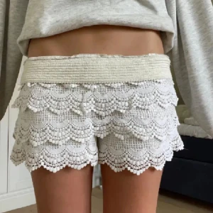 Vita spetsiga shorts med volang - Söker dessa superfina vita shorts med flera lager spets och volangdetaljer. De har en bred, elastisk midja och ett unikt mönster med små bollar längs kanterna. Perfekta för sommarens varma dagar och ger en söt och trendig look.