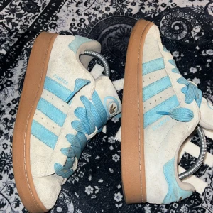 Adidas Campus - Snygga Adidas Campus sneakers i beige mocka med ljusblå detaljer och klassiska tre ränder. Skorna har breda ljusblå skosnören, gummisula i brunt och retro Adidas-logga på plösen. Perfekta för dig som gillar streetstyle och vill sticka ut.