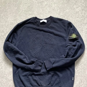 Svart Stone Island sweatshirt - Säljer en svart sweatshirt från Stone Island med stort broderat logomärke på bröstet och klassisk patch på ärmen. Tröjan har rund hals, långa ärmar och är gjord i mjuk bomull. Perfekt för dig som gillar streetwear och vill ha något stilrent.