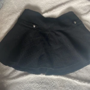 Svart kortkjol från H&M, stl 42 - Snygg svart kortkjol från H&M i storlek 42. Kjolen har en lätt  dekorativa knappar framtill och en lite nopprig yta. Perfekt att matcha med en stickad tröja eller crop top för en trendig look. Är lite nopprig men skulle säga att materialet ska se ut så 