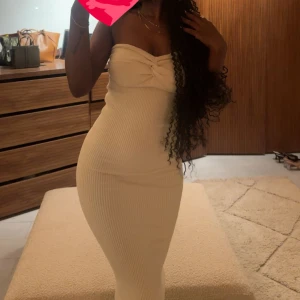 Beige ribbad axelbandslös långklänning - Supersnygg beige ribbad långklänning med axelbandslös design och twistad detalj vid bysten. Klänningen sitter tight och framhäver figuren, perfekt för dig som vill ha en stilren och trendig look.