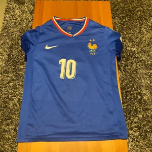 Frankrike Mbappé #10 Nike fotbollströja XS - Blå officiell Frankrike fotbollströja från Nike med Mbappé och nummer 10 på ryggen. Tröjan har korta ärmar, broderad tupp-logga, två stjärnor, och röd-vit-blå krage. Materialet är lätt och andas, perfekt för match eller träning. UEFA Foundation-märke på ärmen.