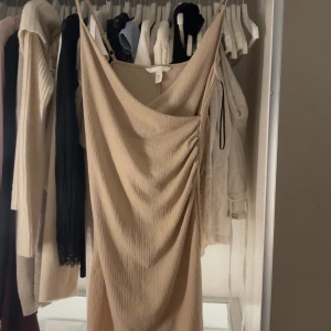 Beige omlottklänning från H&M XS - Säljer en beige kortklänning från H&M i storlek XS. Klänningen har tunna axelband, omlott-drapering och snygg rynkdetalj på sidan. Materialet är strukturerat och känns lätt mot huden, perfekt för varma dagar.