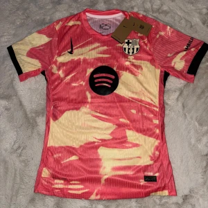 Barcelona x Nike fotbollströja - Unik Barcelona fotbollströja från Nike med coolt rosa/rött och gult mönster, svart Nike-logga och FCB-emblem på bröstet. Spotify-logga framtill och UNHCR-tryck bak. Tillverkad i lätt, ventilerande funktionsmaterial. Perfekt för dig som vill gå runt med bra stil.