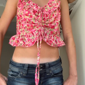 Blommig croppad linnetopp med volang - Supersöt croppad linnetopp från New yorker med blommigt mönster i rosa, rött och grönt. Toppen har volangdetaljer och snörning framtill som ger en extra feminin vibe. Perfekt för varma dagar och somriga outfits! Den är i s men käns lite som xs man kan också justera hur lång och kort toppen kan bli säljs inte längre 💞