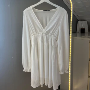 Vit kortklänning från lager 157 med v-ringning - Superfin vit kortklänning från lager 157 med v-ringning och smockad midja. Långa ärmar med lätt puff och resår vid ärmslut. Tunt, flowy tyg som ger en luftig känsla. Använd en gång!💞
