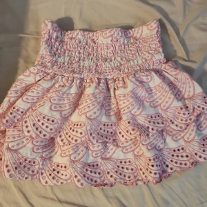 Rosa  kortkjol från Kappahl - Supersöt kortkjol från Kappahl i vitt med rosa broderade detaljer och volangkant. Kjolen har bred smock i midjan för en bekväm passform och ett unikt mönster som verkligen sticker ut. Kjolen är väldigt stretchig 