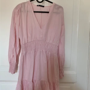 Ljusrosa klänning från Zara - Supersöt ljusrosa klänning från Zara med v-ringning, smock i midjan och långa ärmar med smock vid ärmslut. Klänningen har en flowy passform och är perfekt för vår och sommar. Tillverkad i ett mjukt bomullsliknande material. Köparen betalar frakten 