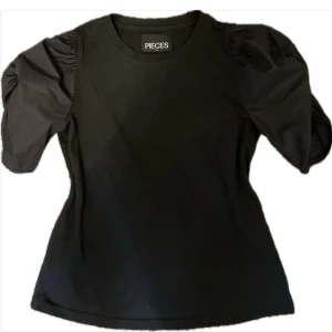 Svart topp med puffärm från Pieces - Svart topp från Pieces i storlek XS med släta lite glansiga puffärmar. Perfekt för dig som vill vara basic men ändå lite finare. 