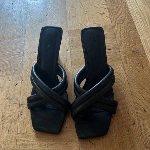 Svarta sandaler med klack från Educe - Stilrena svarta sandaler med fyrkantig tå och korsade, vadderade remmar. Klack för extra höjd och en modern look. Tillverkade i skinnimitation som ger en lyxig känsla. Perfekta för dig som gillar trendiga detaljer och minimalistisk design.