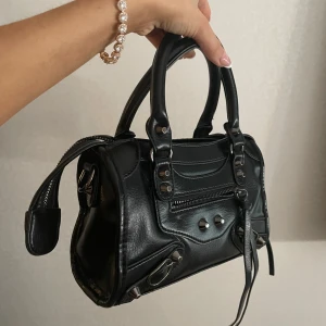 Svart handväska i skinnstil - Cool svart handväska med silvriga metalldetaljer och dragkedja framtill. Väskan har två rejäla handtag och snygga dekorativa nitar. Den är gjord i skinnliknande material och har en urban vibe med detaljer som ger extra edge.