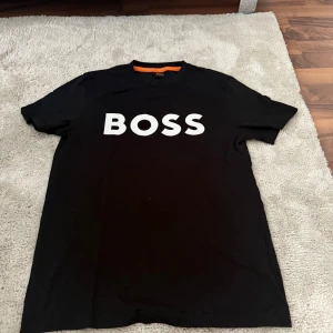 Svart BOSS t-shirt  - Svart t-shirt från BOSS med klassisk passform och stort vitt BOSS-tryck på bröstet. T-shirten har rund halsringning, korta ärmar och är tillverkad i mjuk bomull. Perfekt för dig som gillar stilrena och enkla plagg med tydlig logga.