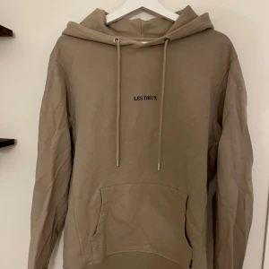 Beige hoodie från Les Deux - Snygg beige hoodie från Les Deux med minimalistisk logga på bröstet. Tröjan har huva med snörning och en stor magficka. Perfekt för dig som gillar en clean och avslappnad stil. 