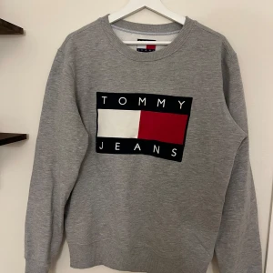 Grå sweatshirt från Tommy Jeans - Snygg grå sweatshirt från Tommy Jeans med stort logotryck i vitt, rött och svart framtill. Klassisk rund halsringning och långa ärmar. Perfekt för en avslappnad och trendig stil.