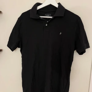 Svart pikétröja från Boomerang  - Klassisk svart pikétröja från Boomerang med liten broderad logga på bröstet. Tröjan har korta ärmar, krage och knäppning med två knappar framtill. Perfekt basic till garderoben och lätt att matcha med allt.