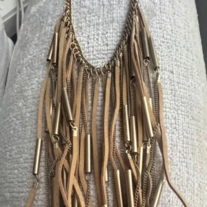 Ny. Beige halsband med fransar från Lindex - Snyggt halsband från Lindex med beige mockaband, guldfärgade kedjor och metallrör som hänger i fransar. Statement-piece som ger boho-vibe och passar perfekt till en enkel t-shirt eller klänning. Justerbar längd med kedja i nacken.