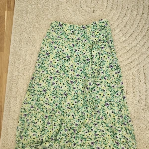 Blommig långkjol från Lindex - Superfin långkjol från Lindex med blommigt mönster i grönt, lila och vitt. Kjolen har elastisk midja och är gjord i ett lätt och luftigt material, perfekt för varma dagar. Snygg att styla med en enkel topp och sneakers.