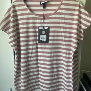 Randig topp i offwhite och rosa från Gozzip - Superfin randig topp från Gozzip i offwhite och rosa. T-shirten har rund halsringning, korta ärmar och en loose passform. Perfekt för dig som gillar en avslappnad och trendig stil. Tillverkad i mjuk bomull som känns skön mot huden. Även kjol till som visas på näst sista bilden. Säljes som ett set. Helt oanvänt. Storlek 44 eller storlek S som det står på lappen vilket motsvar storlek 44 för lite kurvigare kvinnor.