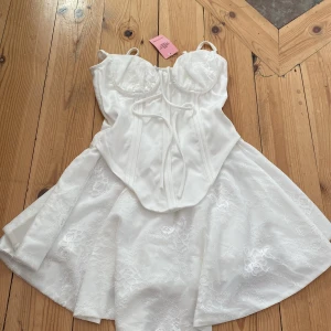 Vit spetsklänning från PrettyLittleThing - Supersöt vit kort klänning med spetsdetaljer och korsettliv från PrettyLittleThing som aldrig har använts. Klänningen har smala axelband, markerad byst, snörning framtill och en utsvängd kjol i spets. Perfekt för dig som vill sticka ut med en romantisk vibe. Den är köpt för 650 kr säljs utan spets armar finns på fler sidor  