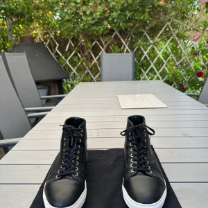 Svarta Versace high-top sneakers - Svarta high-top sneakers från Versus Versace i skinn med vita sulor och guldfärgade öljetter. Sidan pryds av en silverfärgad Versus-medaljong. Klassisk snörning framtill och diskret logga på hälen och sulan. Snygga och stilrena skor med exklusiv känsla. Obs! Med skorna medföljs endast dustbag🙂.skorna säljs även inte längre vilket gör de exklusiva och extremt unika🙌När jag köpte de låg de på runt 5500 kr nypris, mitt pris 2350. Har ni några frågor eller funderingar så är det bara att hojta till🙌 