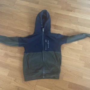 Blågrön hoodiejacka från Sail Racing XS - Snygg tvåfärgad hoodiejacka från Sail Racing i svart och grönt. Jackan har hel dragkedja, huva med snörning och en bröstficka med dragkedja. Perfekt för dig som gillar sportig och avslappnad stil.