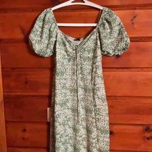 Grön blommig klänning Gina Tricot XS - Supersöt grön klänning från Gina Tricot i storlek XS med vita blommor, puffärmar och snörning framtill. Klänningen har v-ringning och är perfekt för sommarens alla tillfällen. Lätt och luftig modell med fin passform.