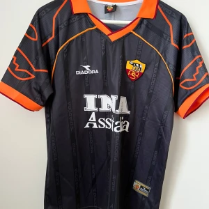 AS Roma Totti Diadora fotbollströja - AS Roma matchtröja med Totti 10 på ryggen. Svart med tunna vertikala ränder och orange detaljer på krage, ärmslut och mönster. Broderat klubbmärke, Diadora-logga och Lega Calcio-patch på ärmen. Tillverkad i lätt polyester, perfekt för fotbollsfans.