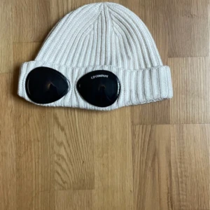Vit C.P. Company mössa - Vit ribbstickad mössa från C.P. Company med ikoniska svarta goggle-linser framtill på uppviket. Klassisk och streetinspirerad design som sticker ut. Perfekt accessoar för dig som vill ha något unikt och trendigt till vintern.